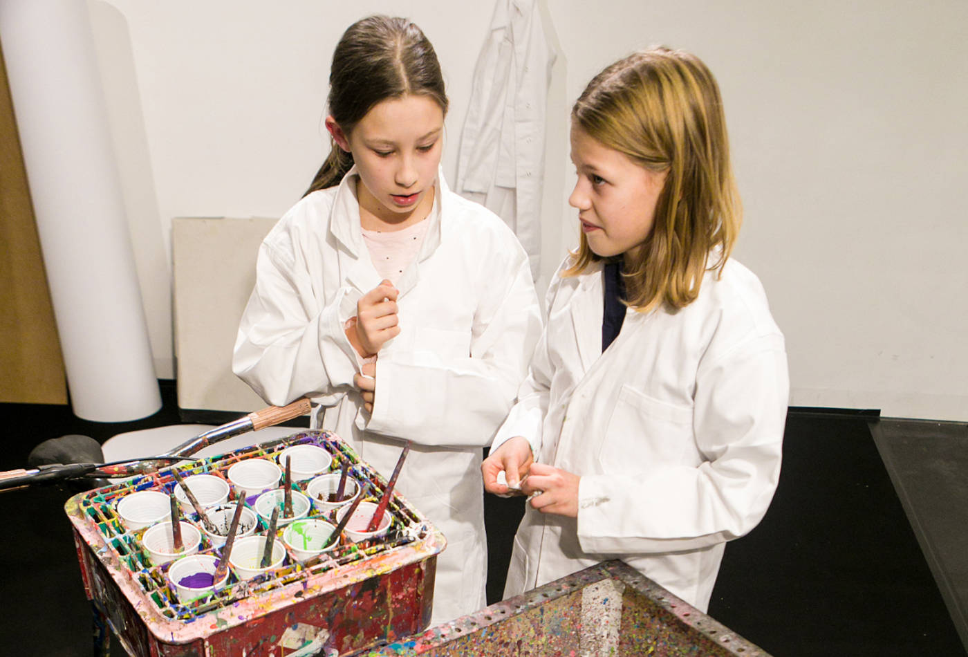 Zwei Mädchen in weißen Kitteln stehen vor einem Farbkasten
